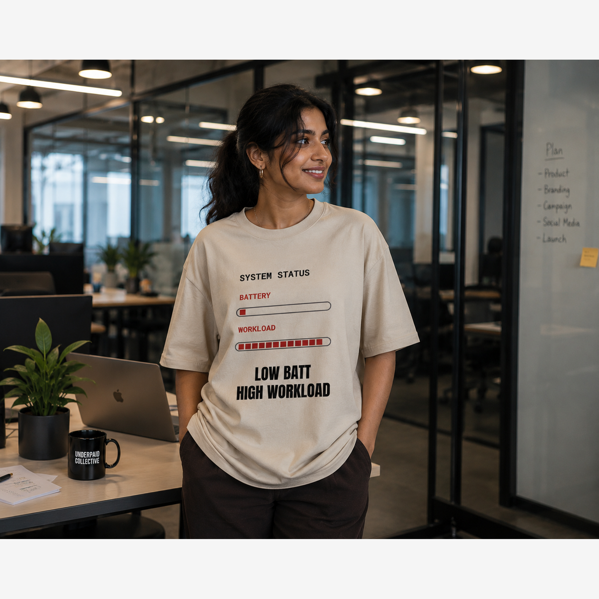 A woman in an office setting wearing a beige graphic t-shirt with a battery status graphic that reads 'LOW BATT HIGH WORKLOAD'.